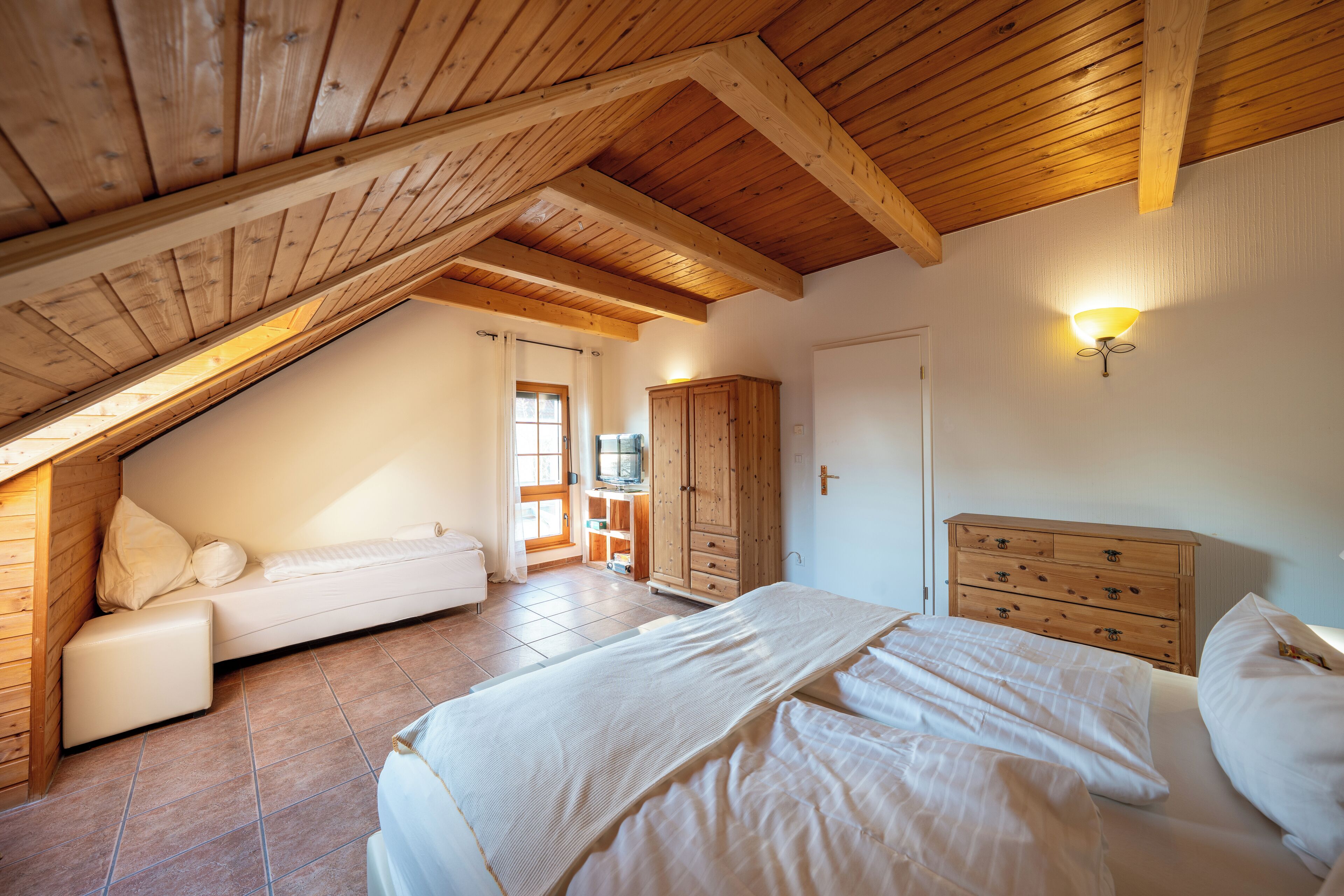 Un dormitorio con techo inclinado, vigas de madera, una cama, un aparador y un televisor en la esquina.