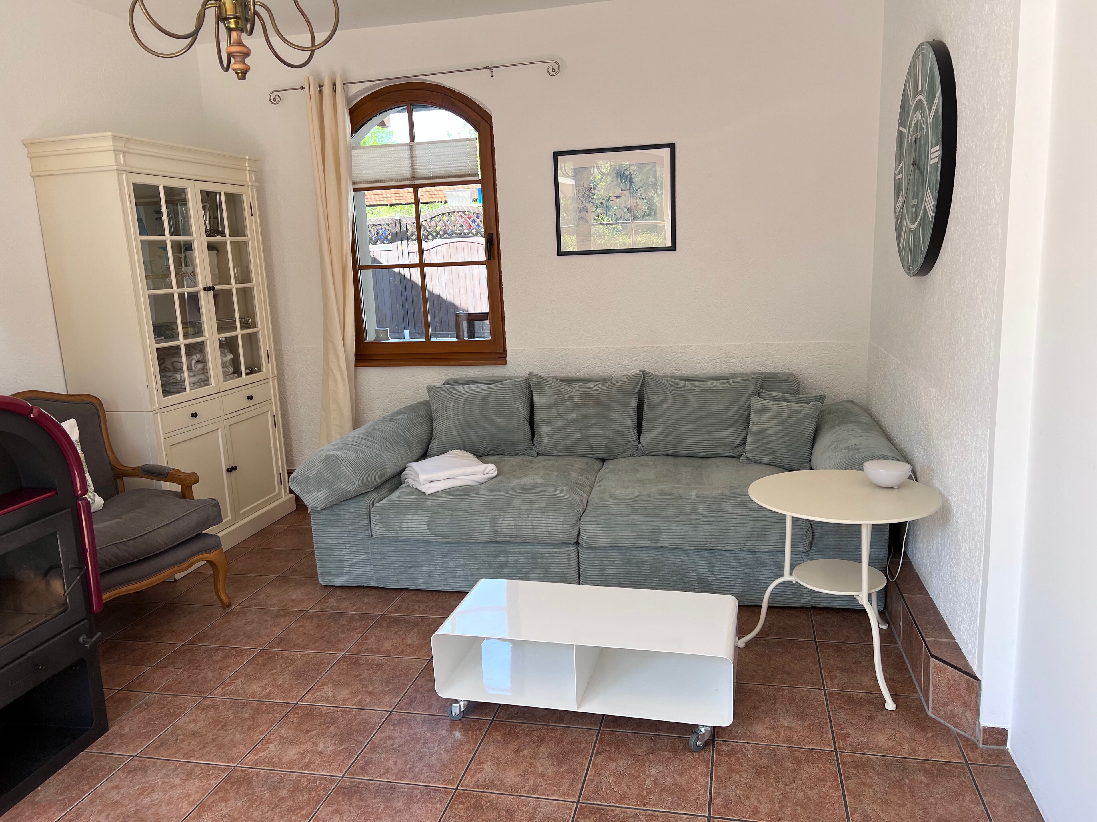 Un salón con un sofá gris, una mesita auxiliar blanca, un mueble con puertas de cristal y una ventana con vistas a una valla.
