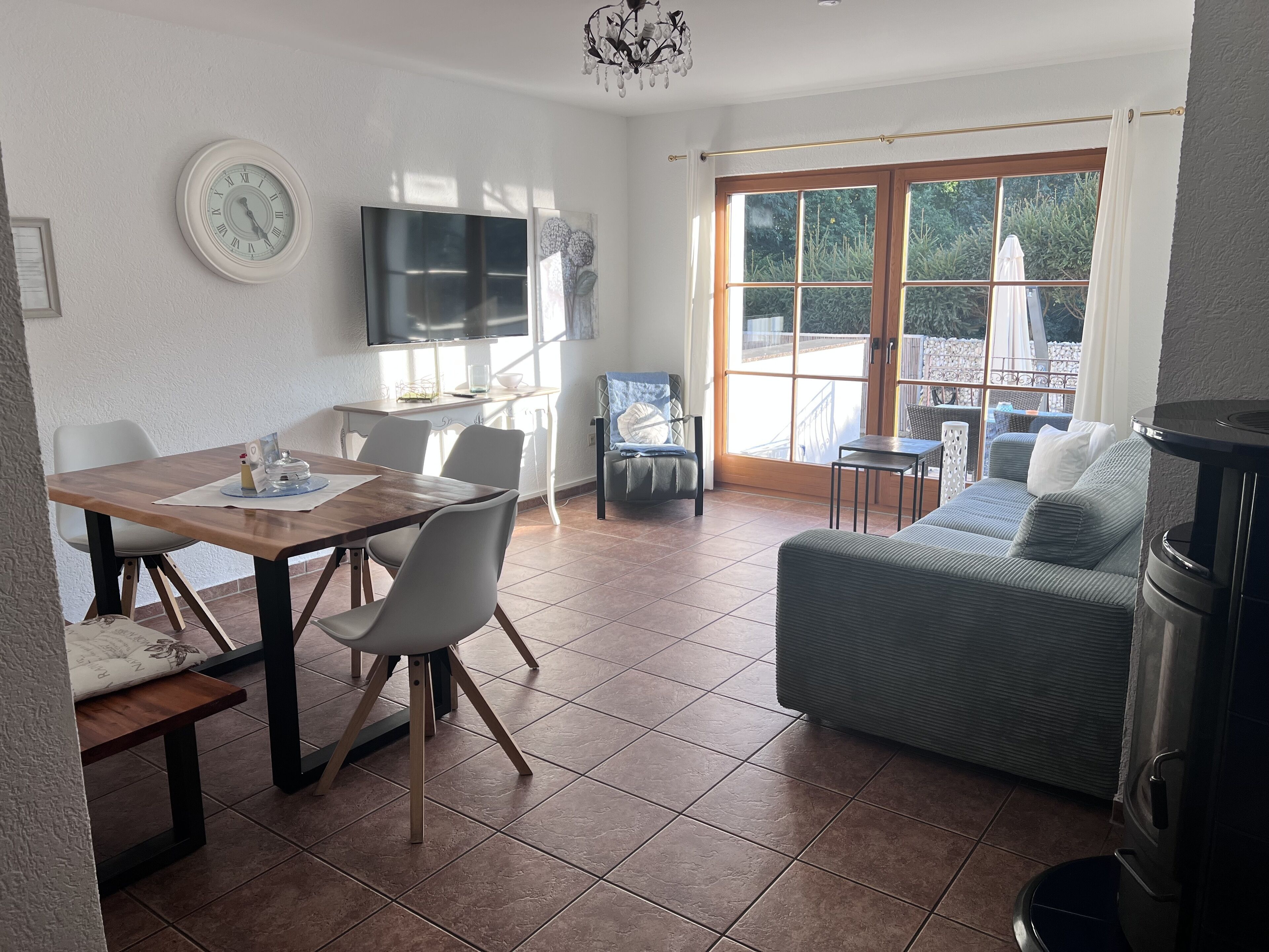 A dining area with a wooden table and chairs, a sofa, a TV, and a large window with a view of greenery.