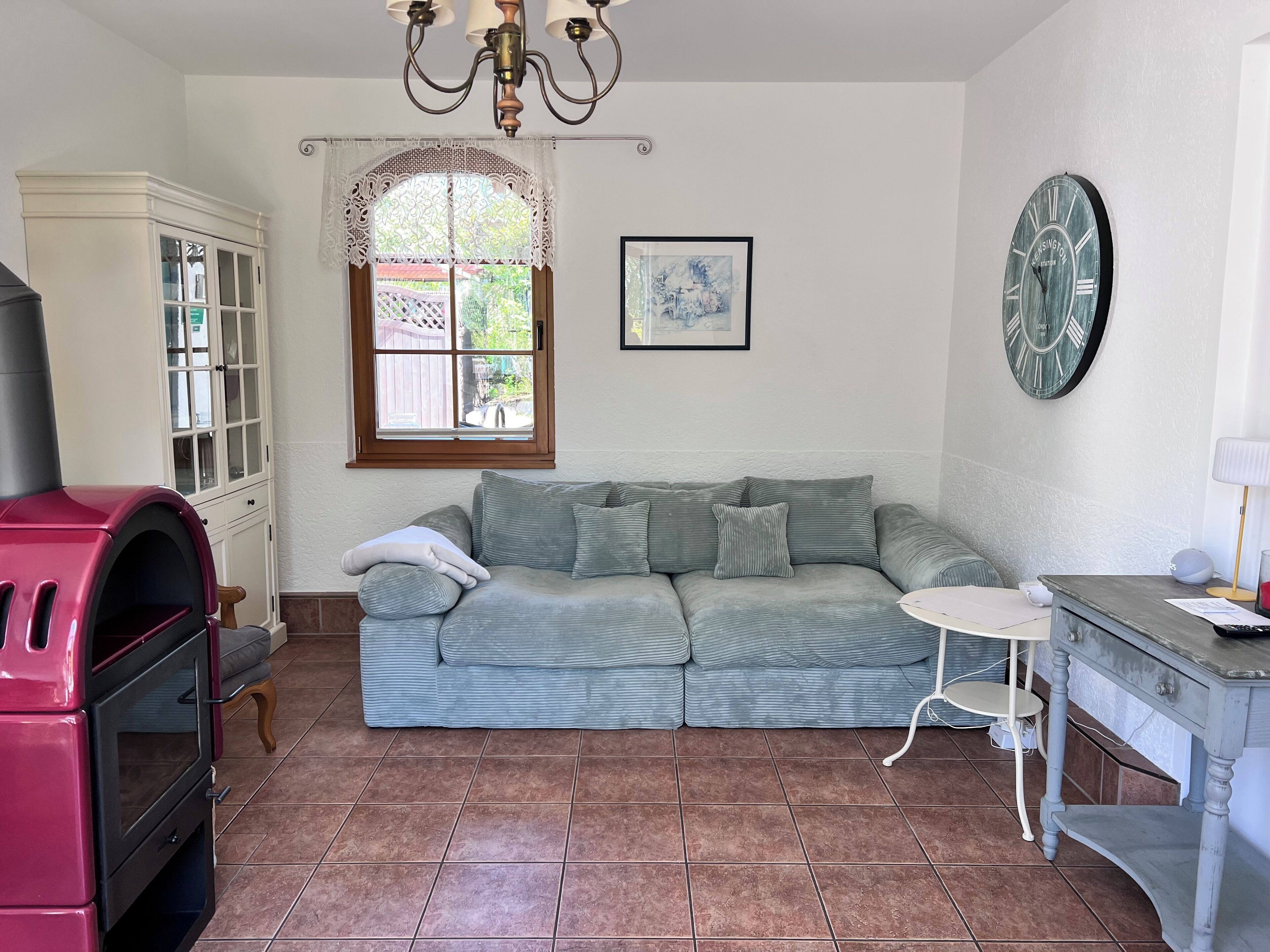 A cozy living room with a sectional sofa, a small round table, a built-in oven, and a window with lace curtains.