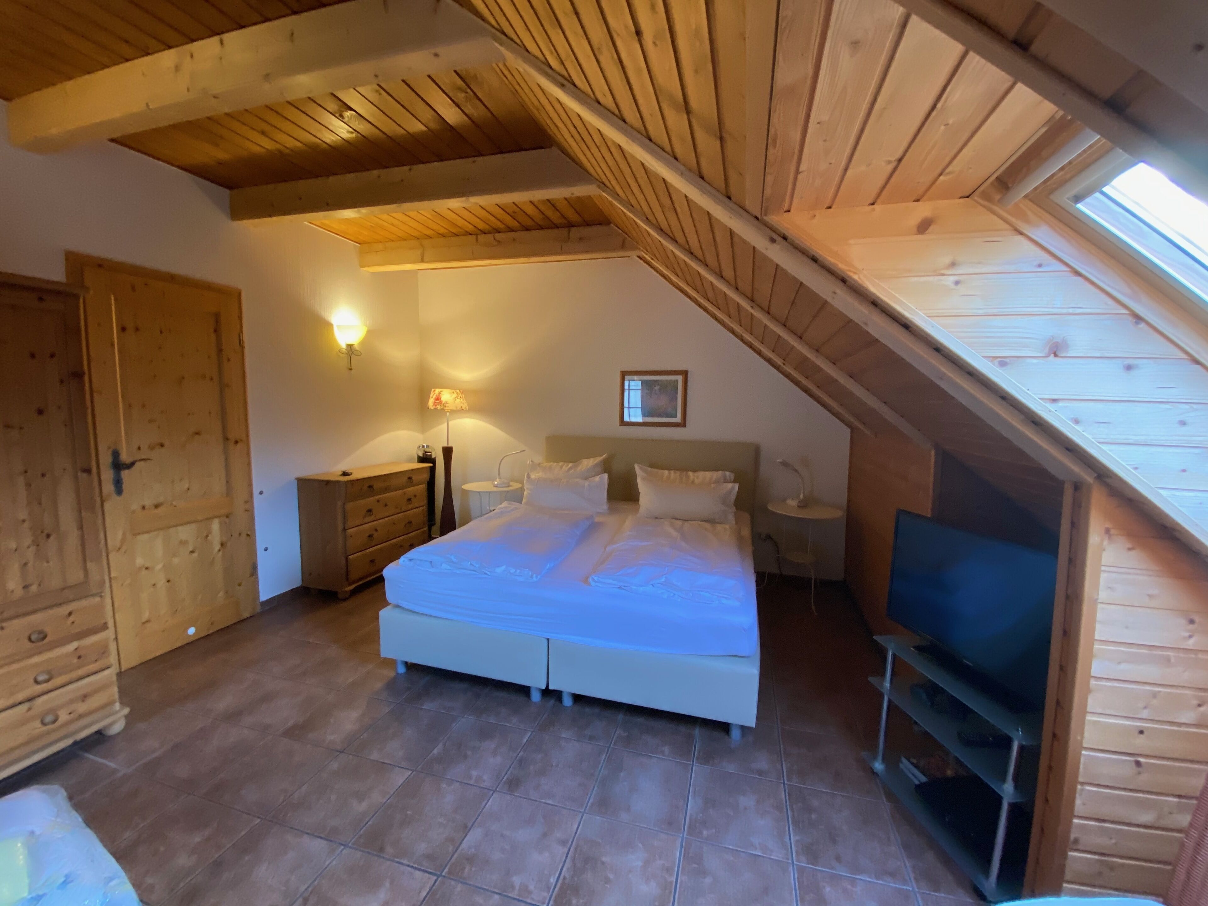 A bedroom with a sloped ceiling, wooden beams, a bed, bedside tables, a lamp, a television, and a door.