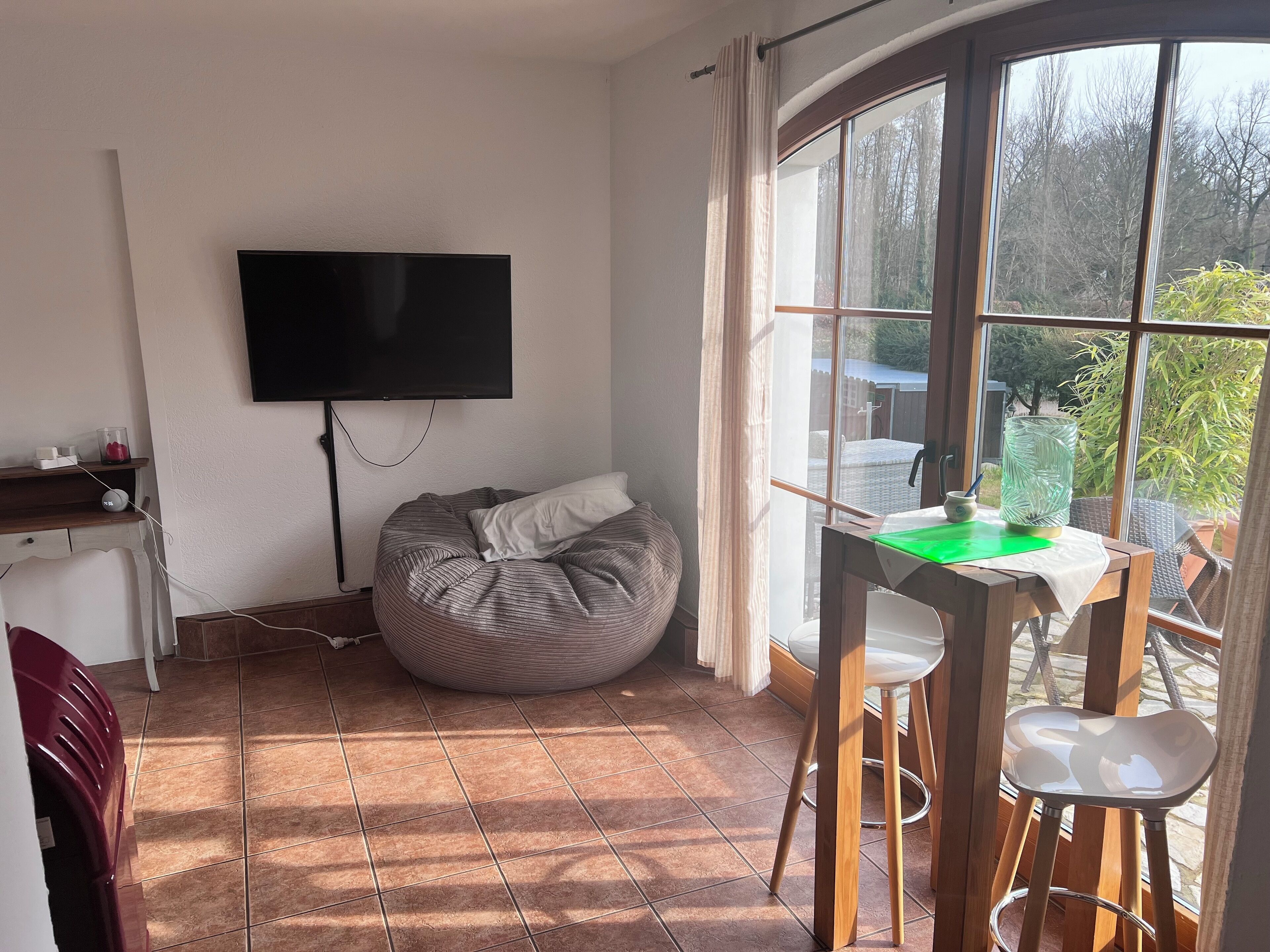 A room with a bed, a bean bag, a TV mounted on the wall, a dining table with chairs, and a large window with a view of trees and a fence.