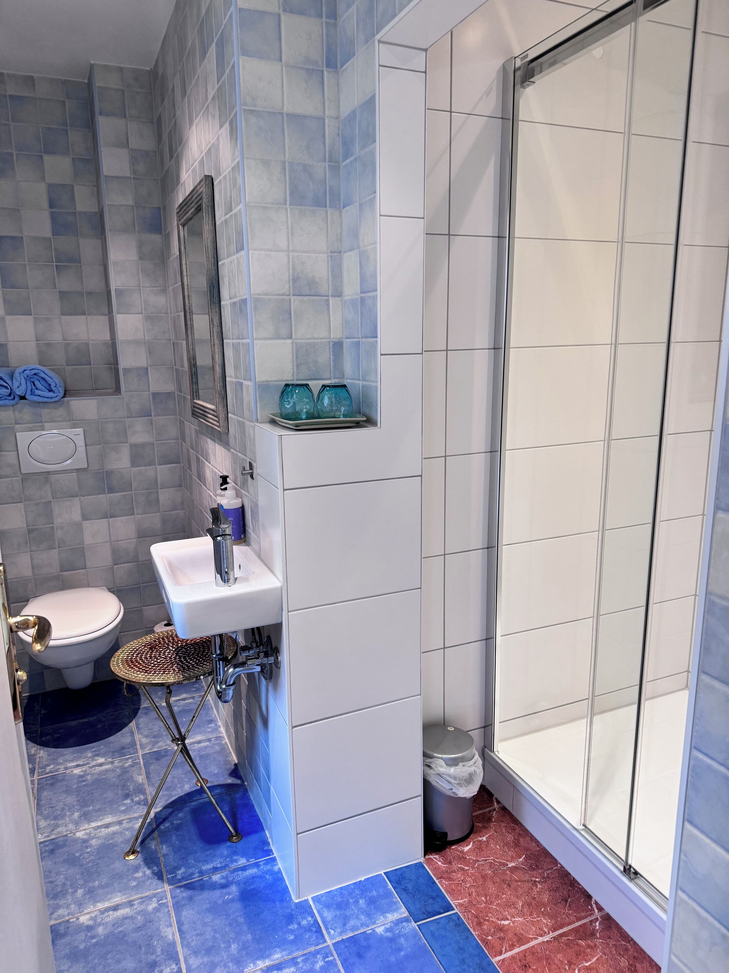 Ein Badezimmer mit weißem Waschbecken, Toilette, Dusche mit Glastür und Fliesen in Blau und Rot.
