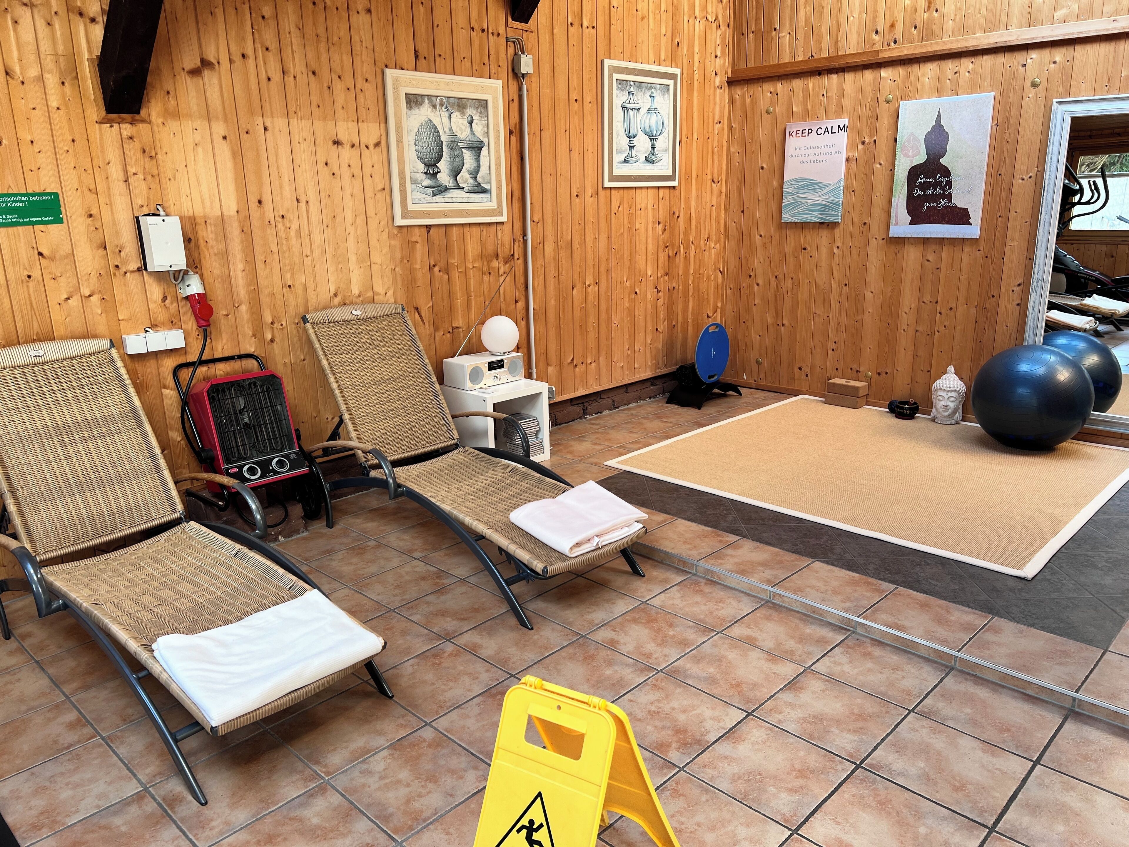 A room with wooden walls, two wicker chairs, a massage table, a massage chair, a small white table with a speaker, and exercise balls.