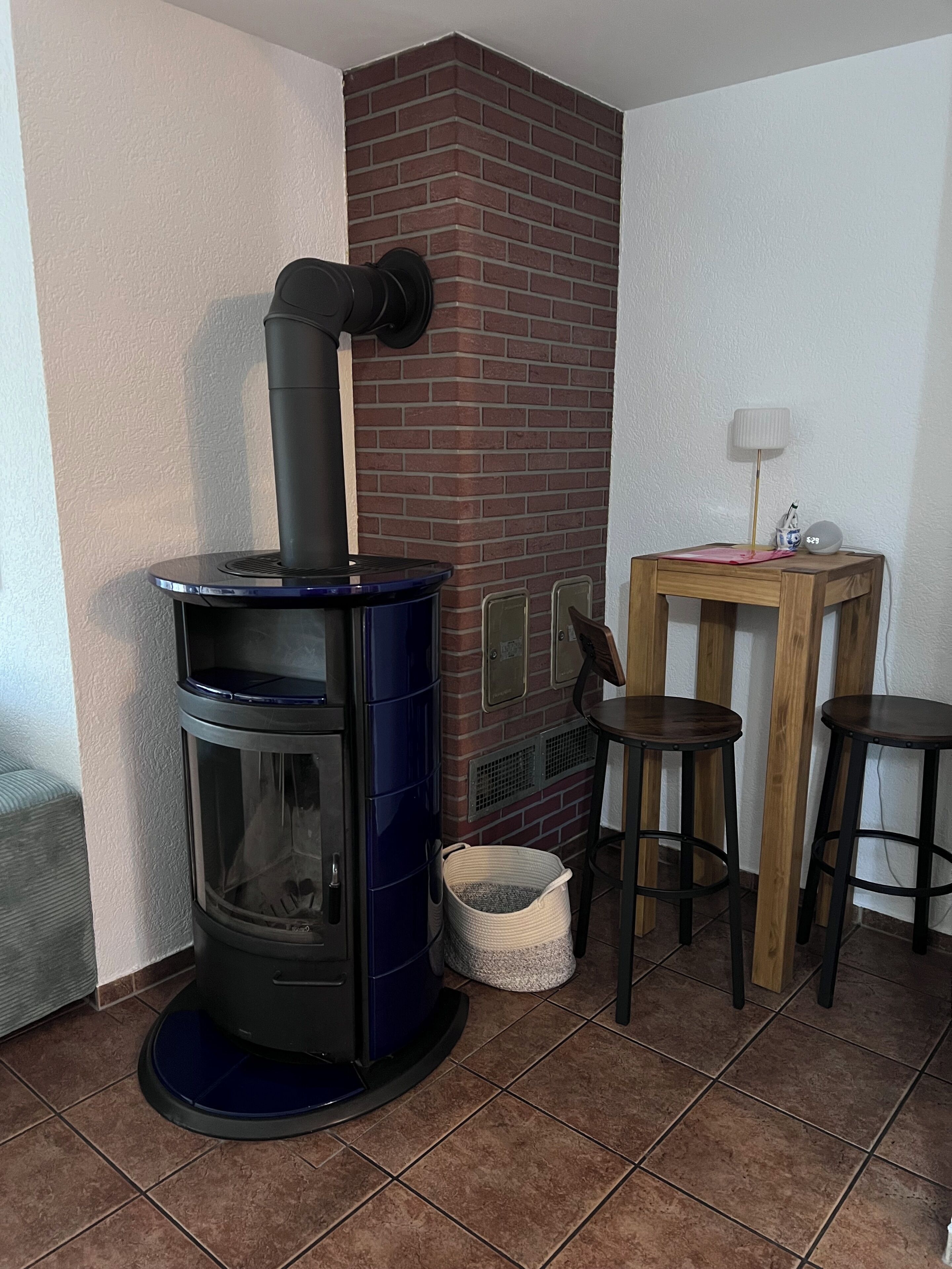 Una habitación con un estufa de leña, una mesita con lámpara y dos taburetes.