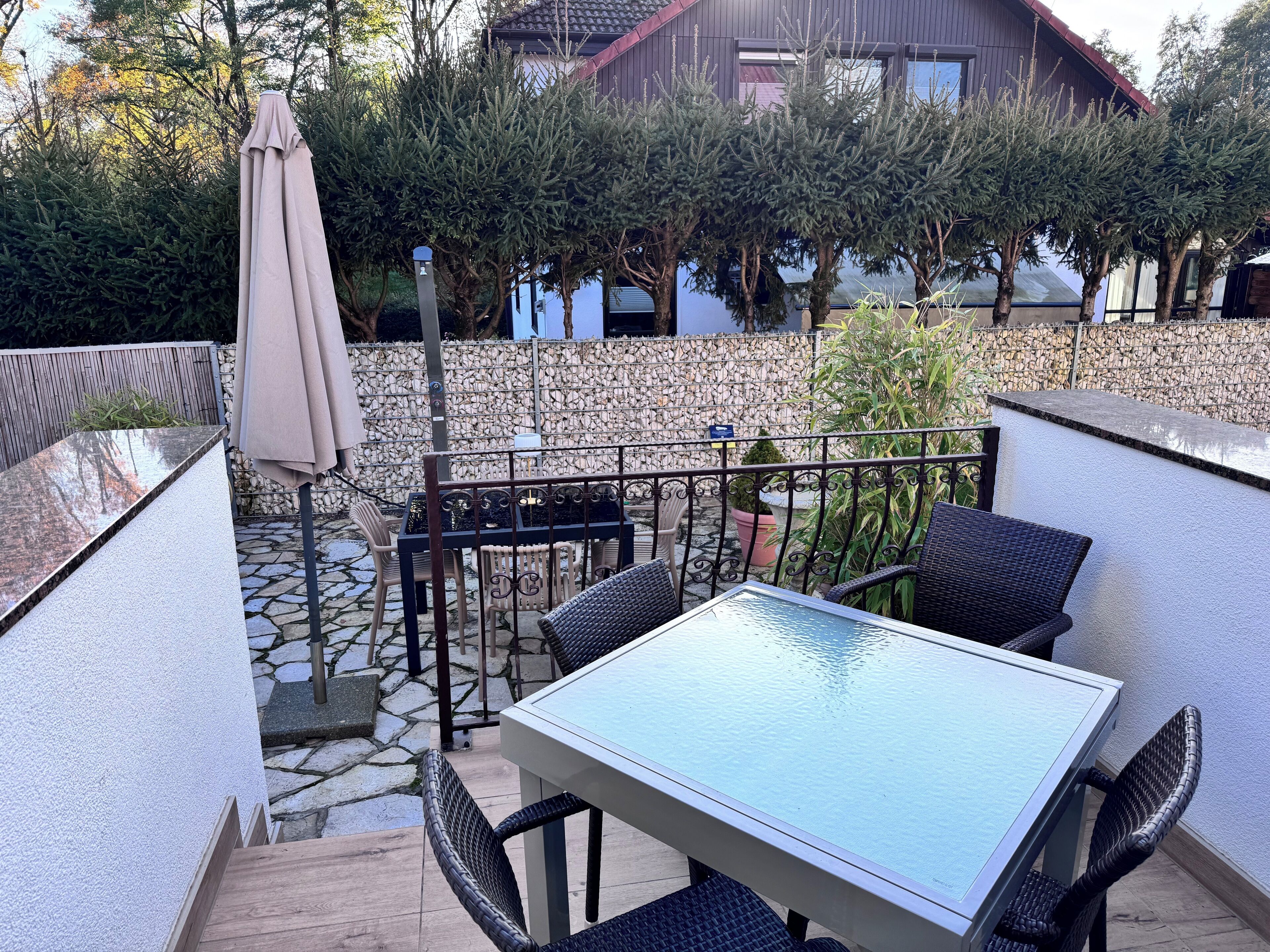Un patio esterno con tavolo in vetro e sedie in vimini, circondato da un muro in pietra e piante in vaso.