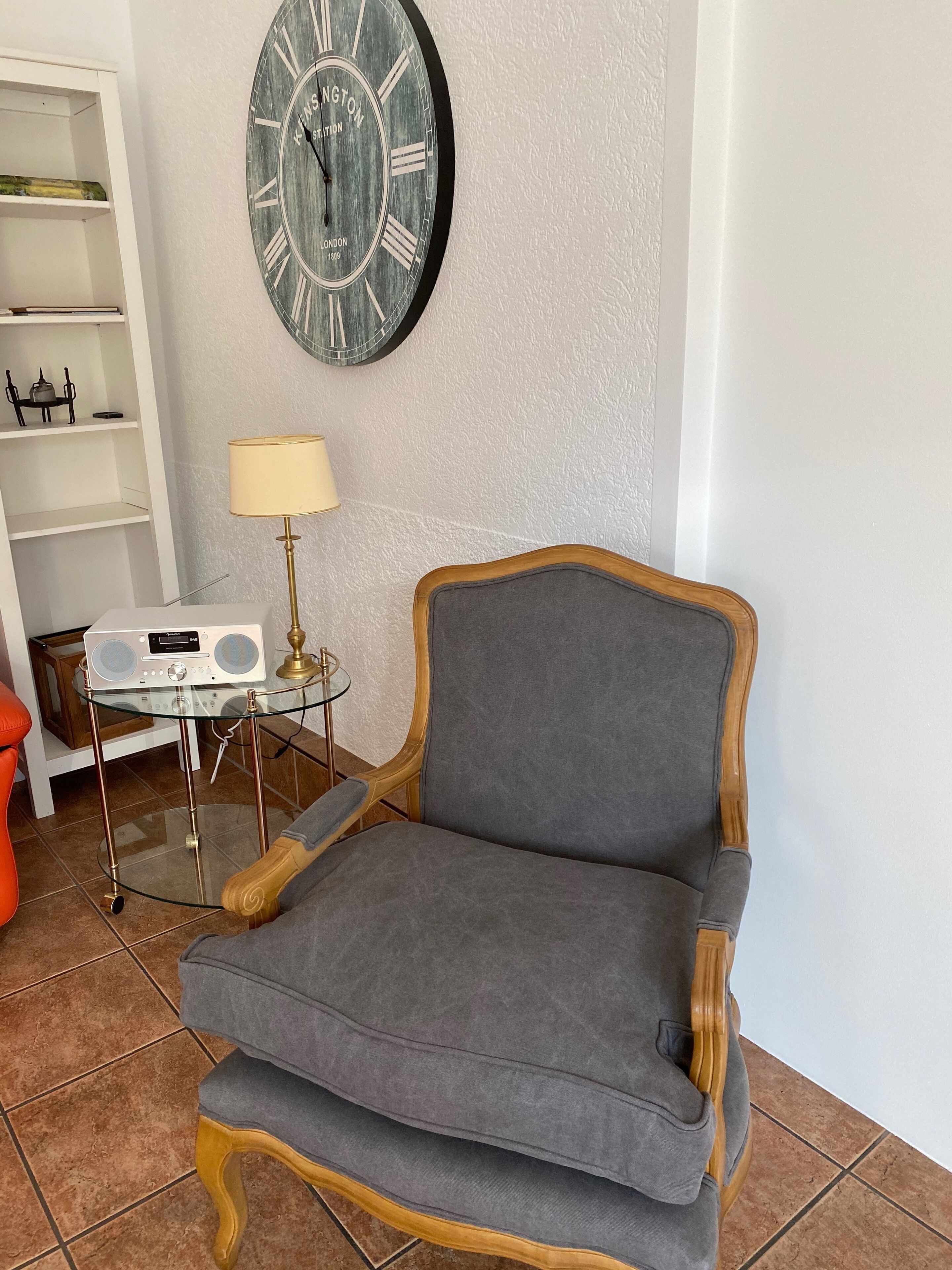 A room with a grey upholstered armchair, a round wall clock, a small table with a radio, and a lamp.