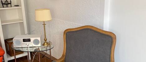 A room with a grey upholstered armchair, a round wall clock, a small table with a radio, and a lamp.