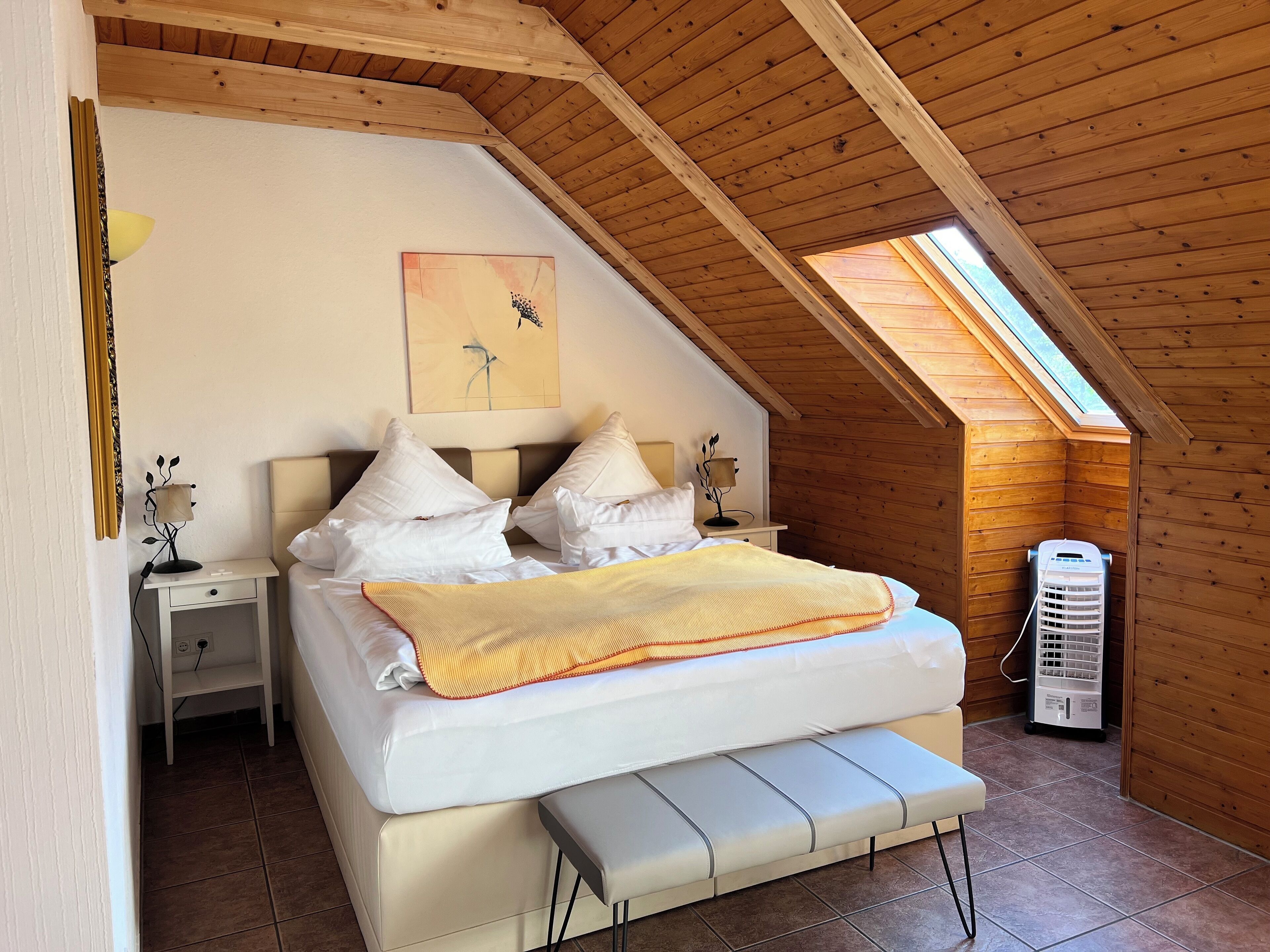 A bedroom with a sloped ceiling, a bed with white bedding, a bench, a bedside table with a lamp, a wall-mounted artwork, and a heater.