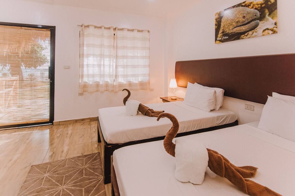 Une chambre d’hôtel avec deux lits, une fenêtre ornée de rideaux légers, un tableau représentant une baleine accroché au mur et un tapis à motifs géométriques.