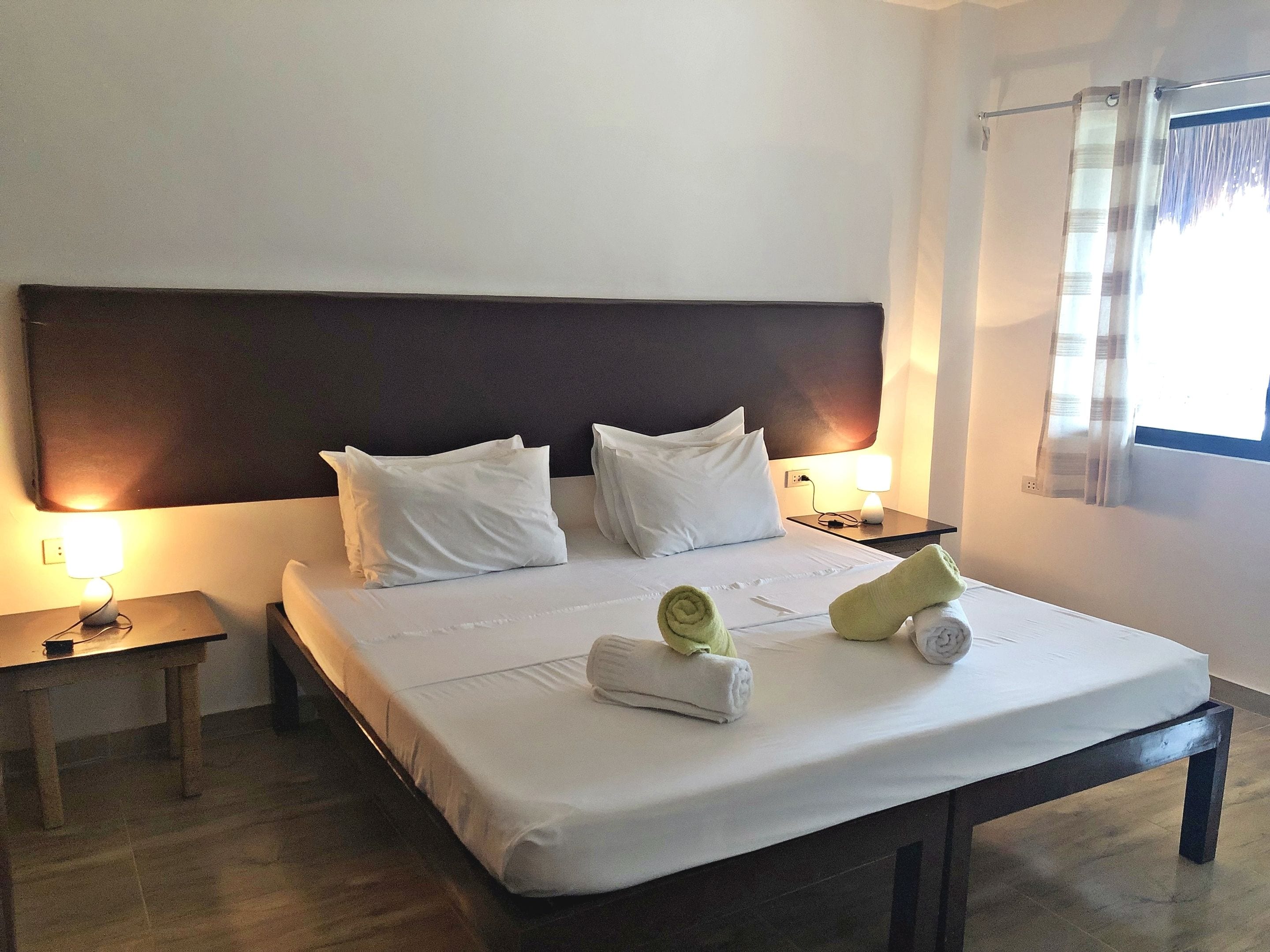 Une chambre d’hôtel avec un grand lit, deux tables de chevet, une petite table et une fenêtre avec des rideaux.