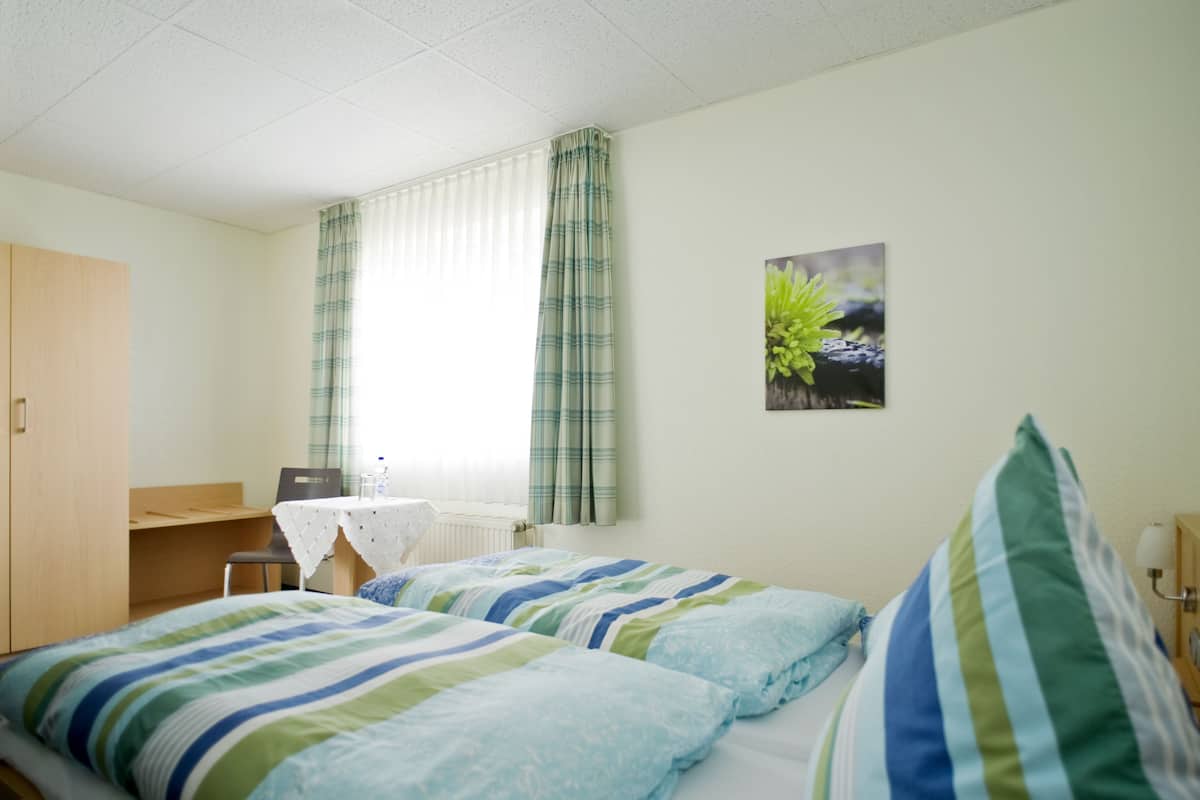 A hotel room with two beds, a desk, a chair, a radiator, and a window with curtains.