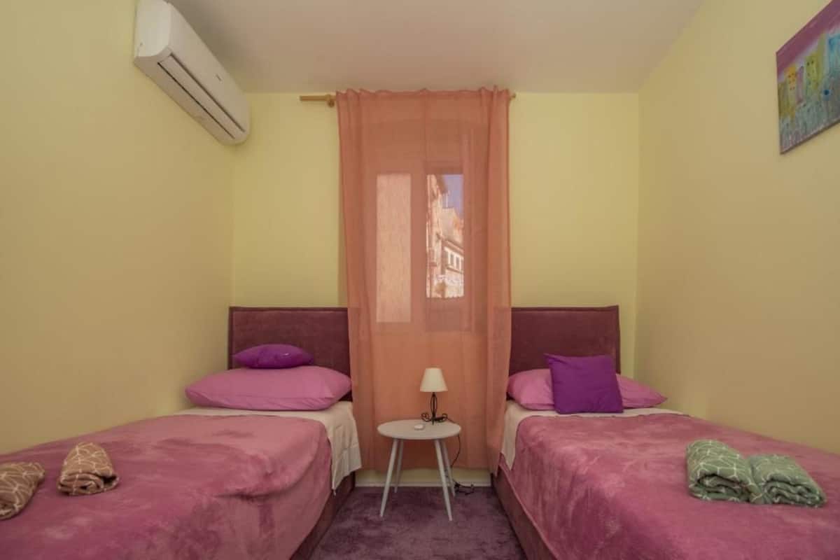 Une petite chambre agréable, dotée de deux lits, d’une petite table avec une lampe et d’un appareil de climatisation.