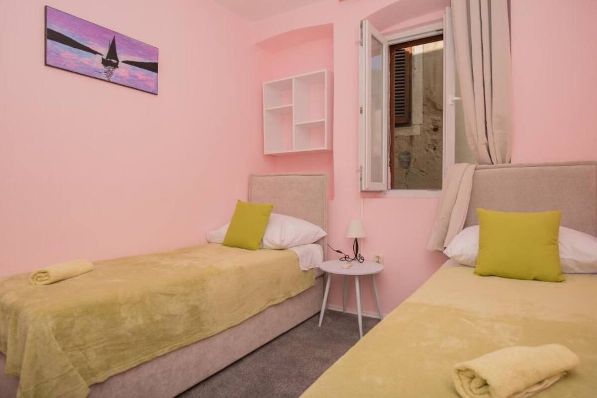 Une chambre rose avec deux lits, chacun recouvert d’une couverture jaune et agrémenté d’un oreiller. On y trouve une petite table blanche avec une lampe, une fenêtre avec des rideaux et une étagère fixée au mur.
