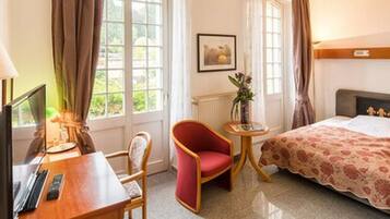 Une chambre d’hôtel avec un lit, un bureau avec un ordinateur, une chaise, une petite table avec un vase et une fenêtre avec des rideaux.