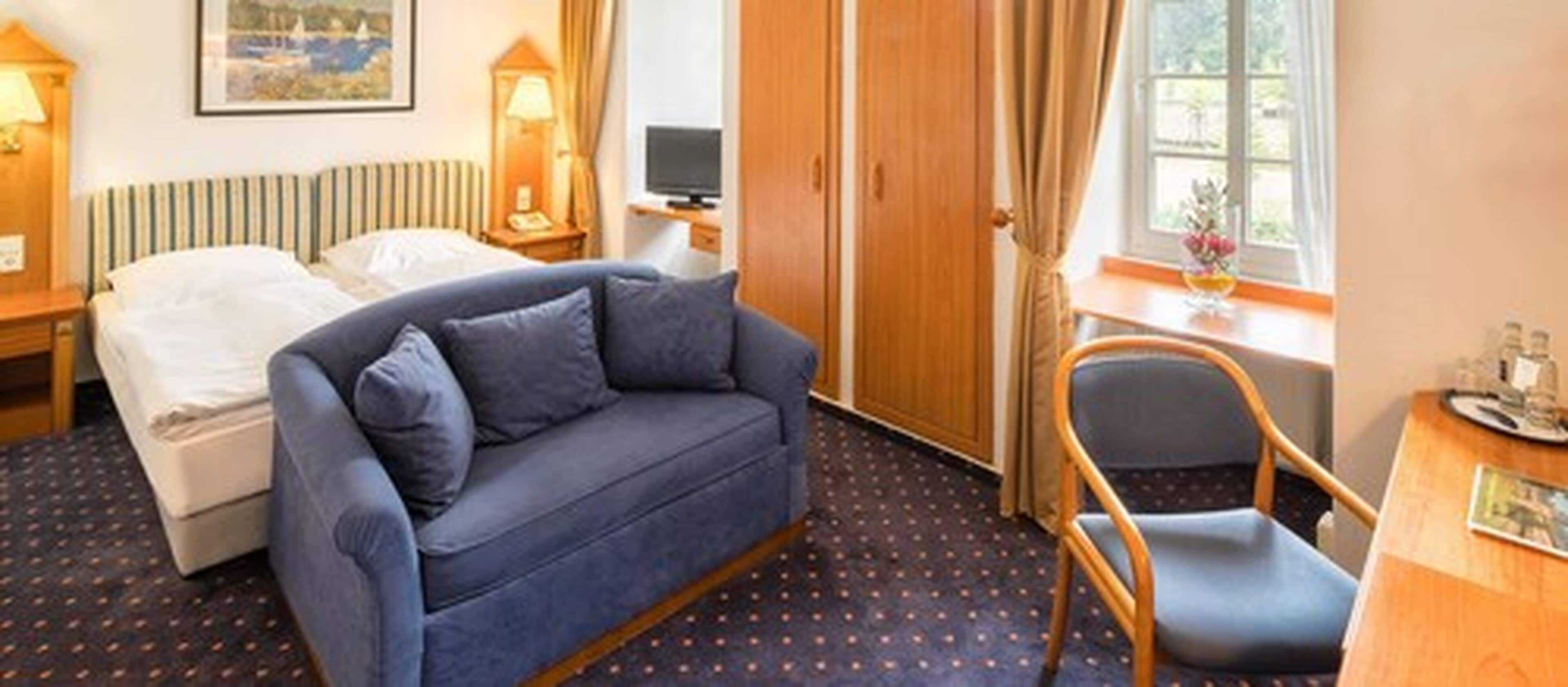 Une chambre d’hôtel avec un lit, un canapé bleu, une chaise en bois, un bureau avec une lampe et une fenêtre avec des rideaux.