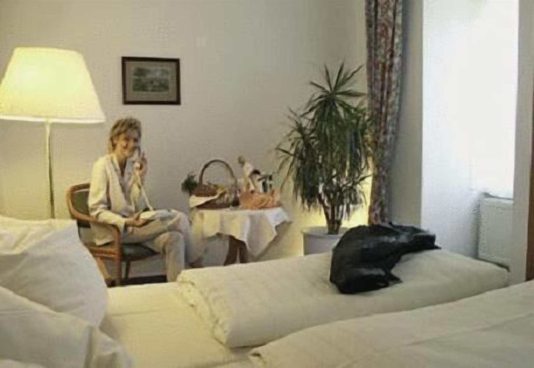 A hotel room with a bed, a chair, a lamp, a table with a plant, and a framed picture on the wall.
