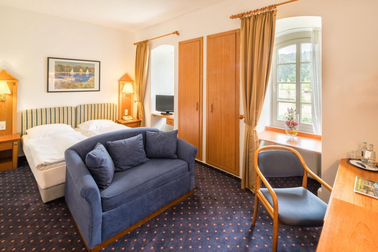 Habitación de hotel con cama, sofá, escritorio, silla, televisión y ventana con cortinas.