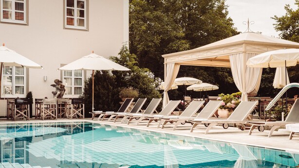 Una piscina con sillas de descanso y sombrillas, rodeada por un edificio con ventanas y árboles.