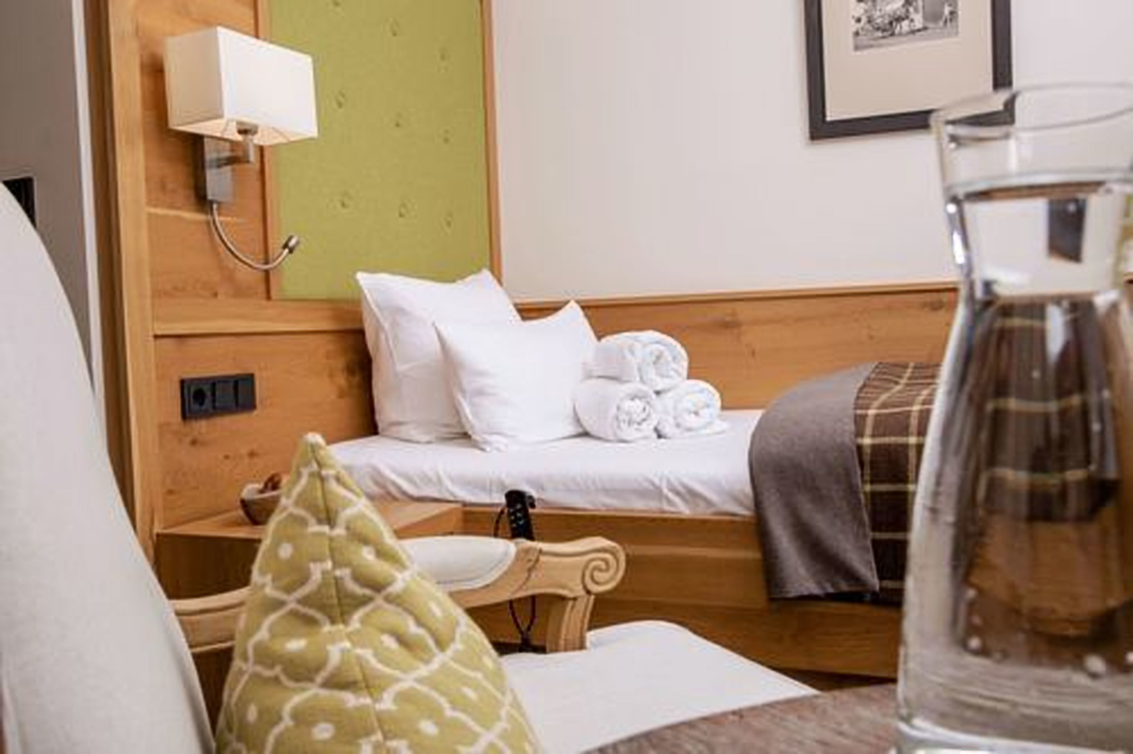 Habitación de hotel con cabecera de madera, cama con sábanas blancas, mesita de noche con lámpara, un cuadro en la pared y un vaso de agua sobre la mesa.