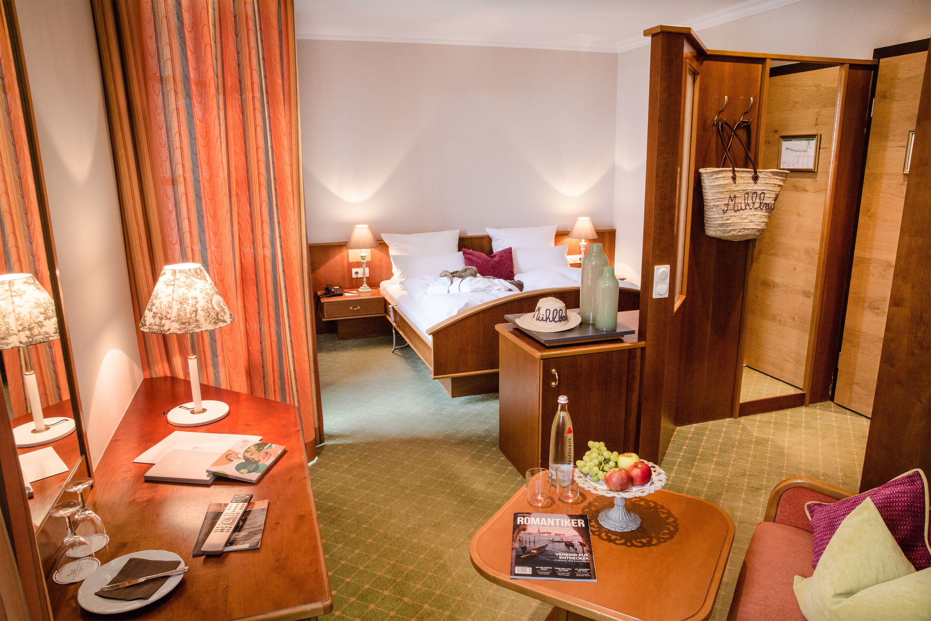 A hotel room with a bed, bedside tables with lamps, a small seating area with a table, and a basket on the wall.