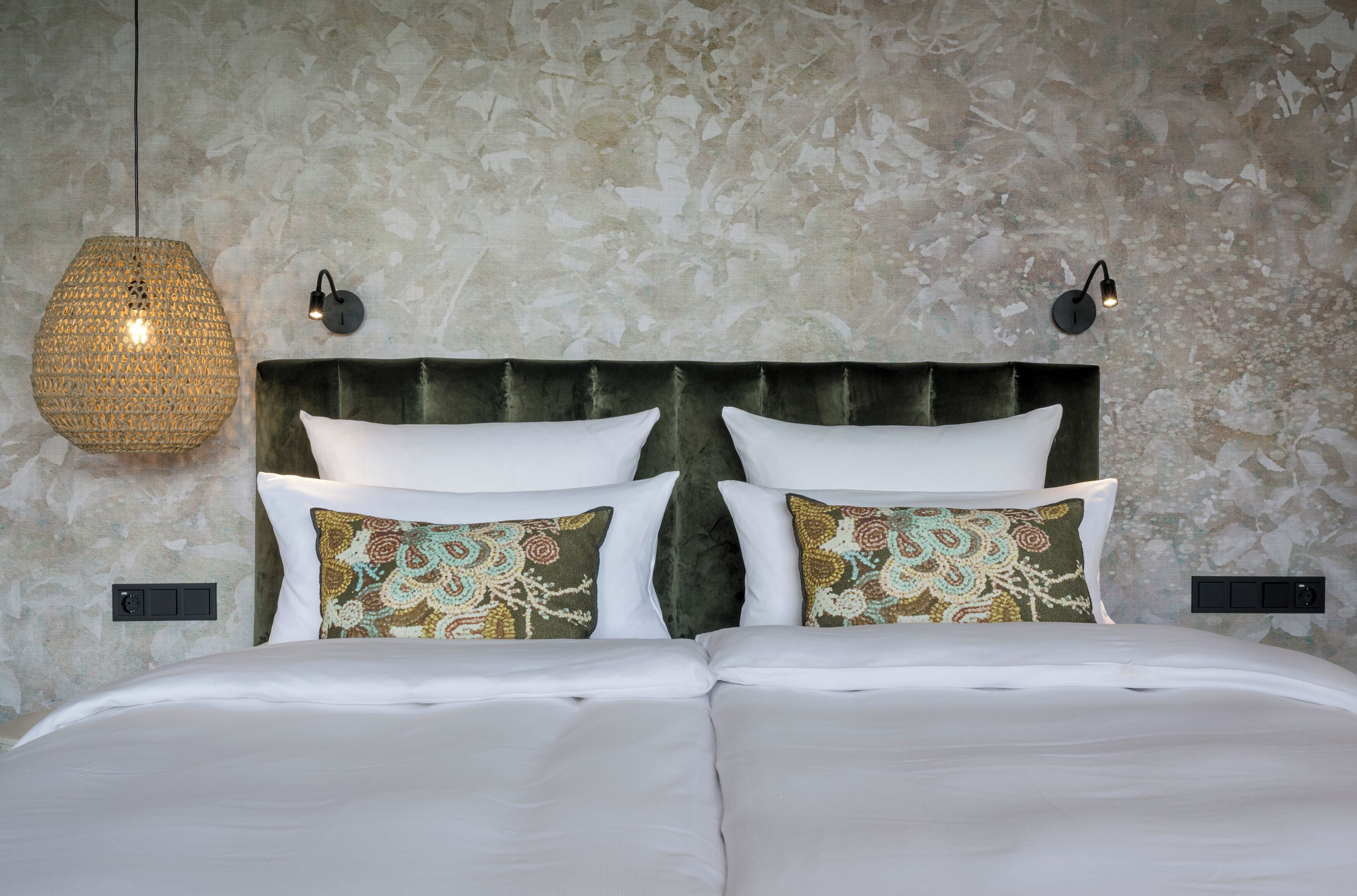 A neatly made bed with white linens and patterned pillows, a textured wall behind, and two wall-mounted lamps.