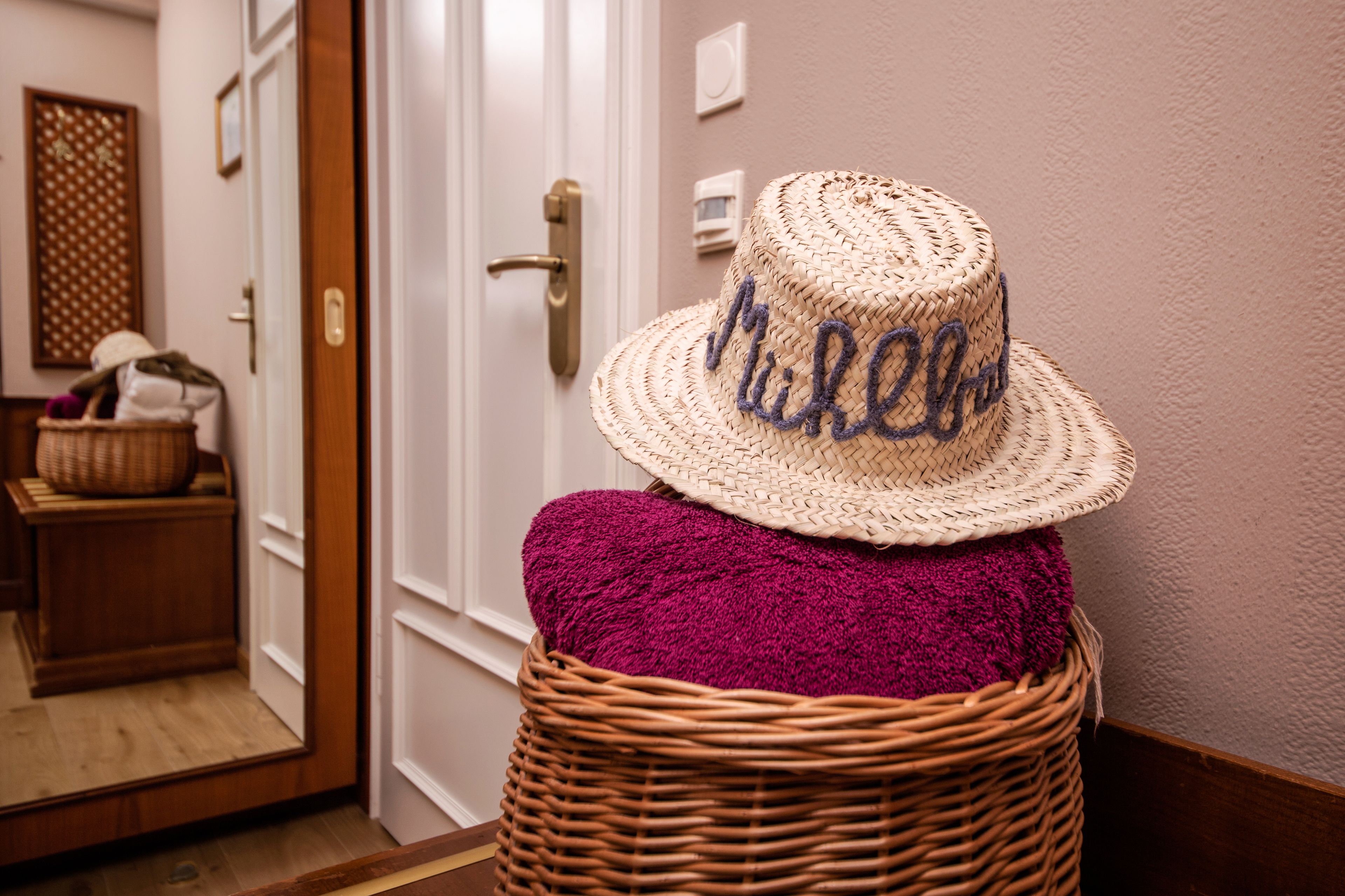 A wicker basket with a straw hat and a purple cushion in a hallway.