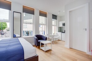 A modern hotel room with a large bed, a seating area with a chair and a small table, a kitchenette, and large windows offering a view of the street. - Chapel Market Serviced Apartments by Concept Apartments (London)