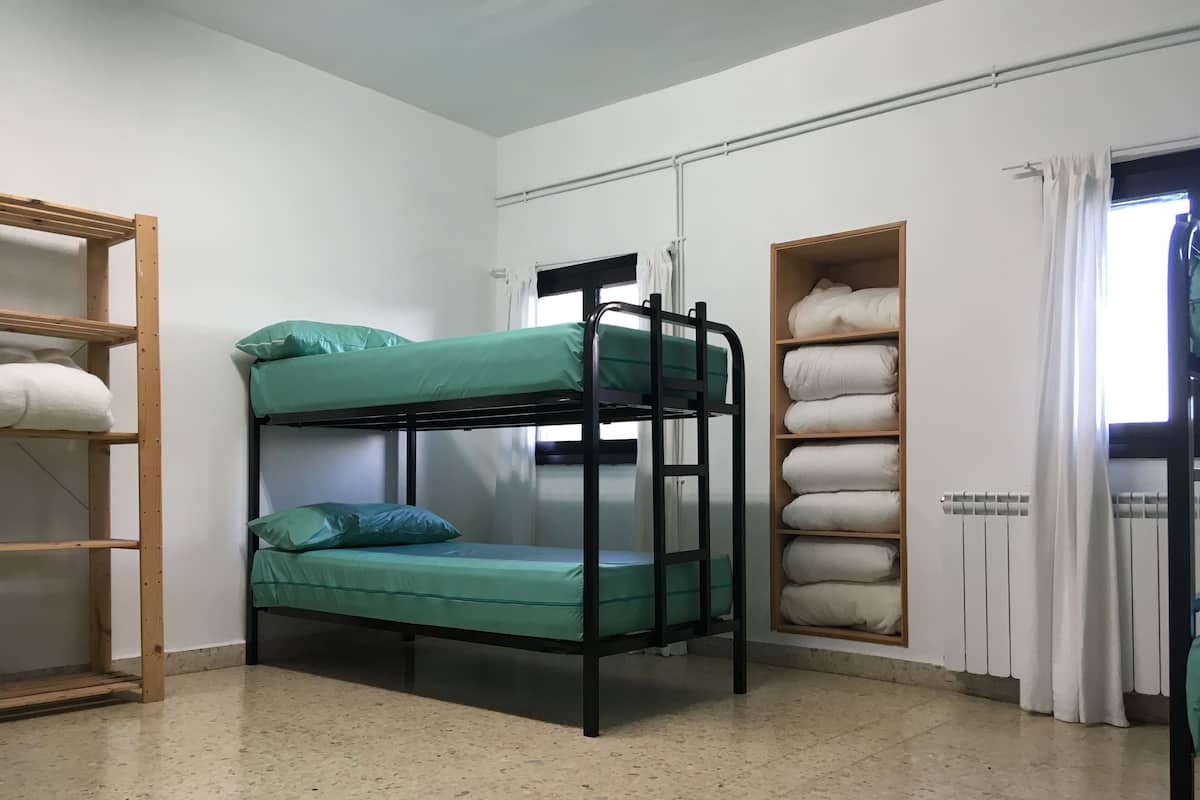 A bunk bed with green bedding, a wooden shelf unit, and a radiator with white curtains.