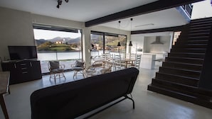 A modern living room with a large window overlooking a scenic landscape, a dining area with wicker chairs, a kitchen with white cabinets, and a dark staircase. - Albergue Abaraxka - Hostel (Azkoitia)
