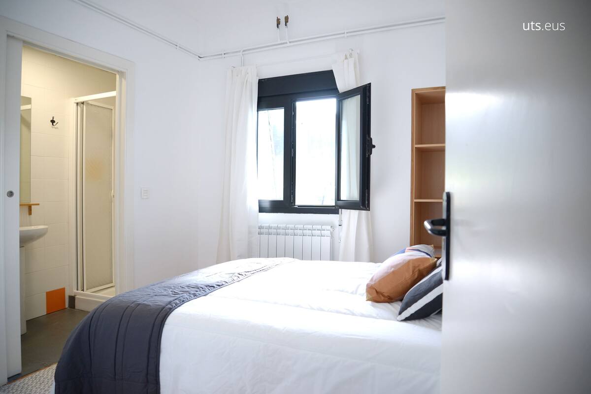 A hotel room with a bed, a window with curtains, a radiator, and a wooden shelf.