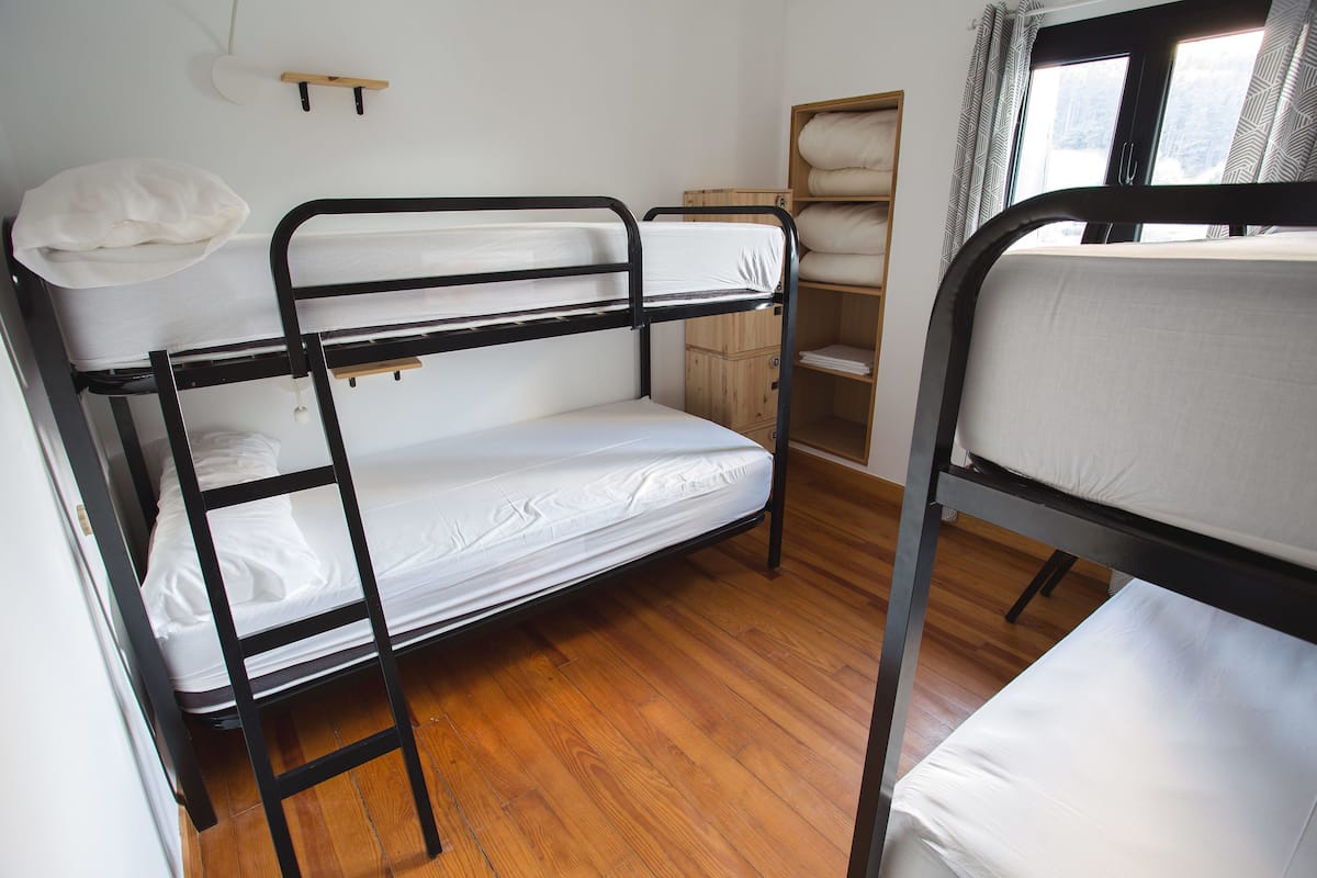 A bunk bed room with a wooden floor, a window with curtains, and a wooden shelf with a white pillow.