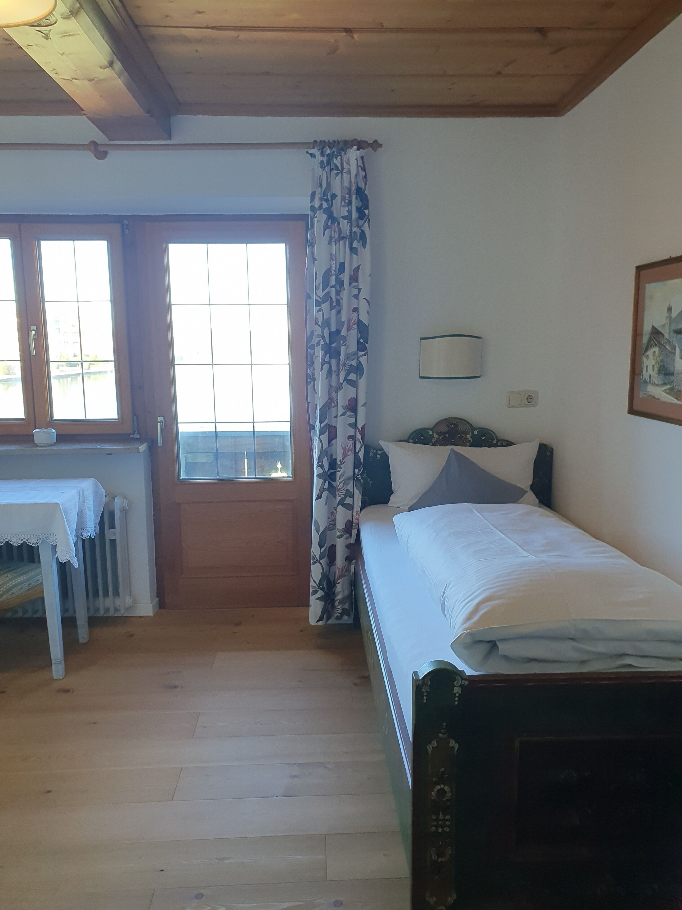 A bedroom with a bed, a table with a lace tablecloth, a window with a view, and a door.