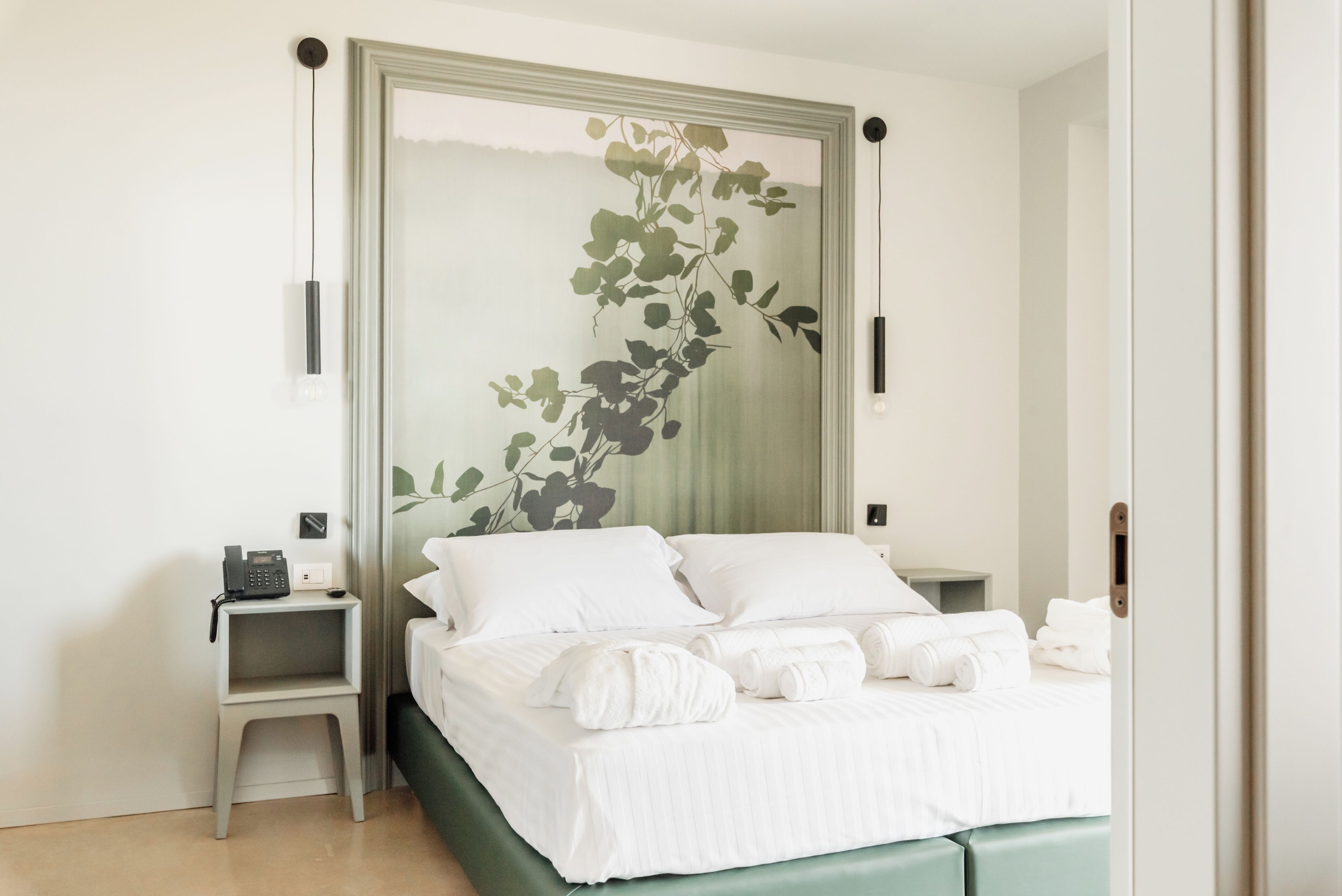 A neatly made bed with white linens and rolled towels, a bedside table with a phone, and a wall-mounted light fixture.