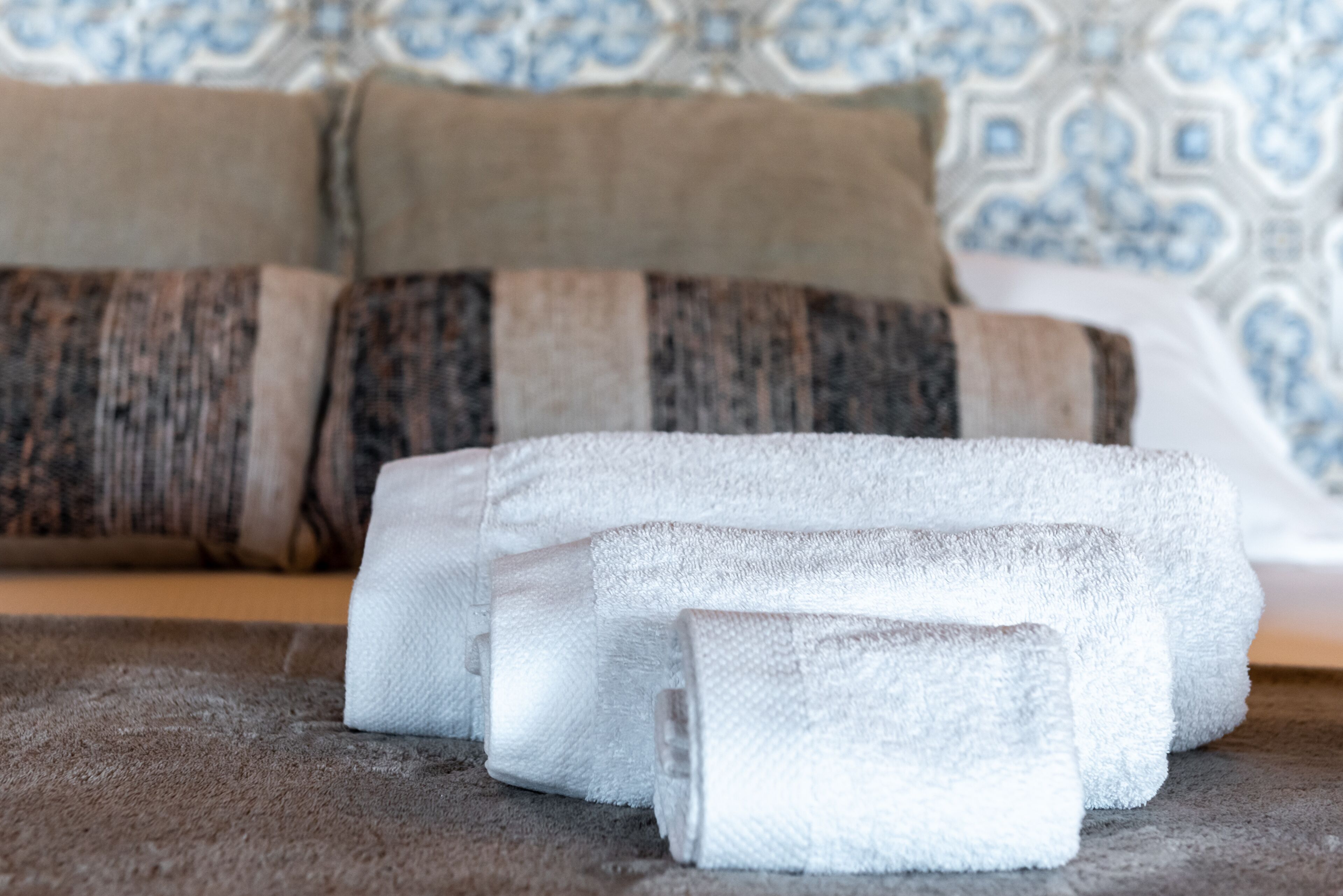 Neatly folded white towels on a textured brown carpet, with a patterned blue and white wall and a brown sofa in the background.