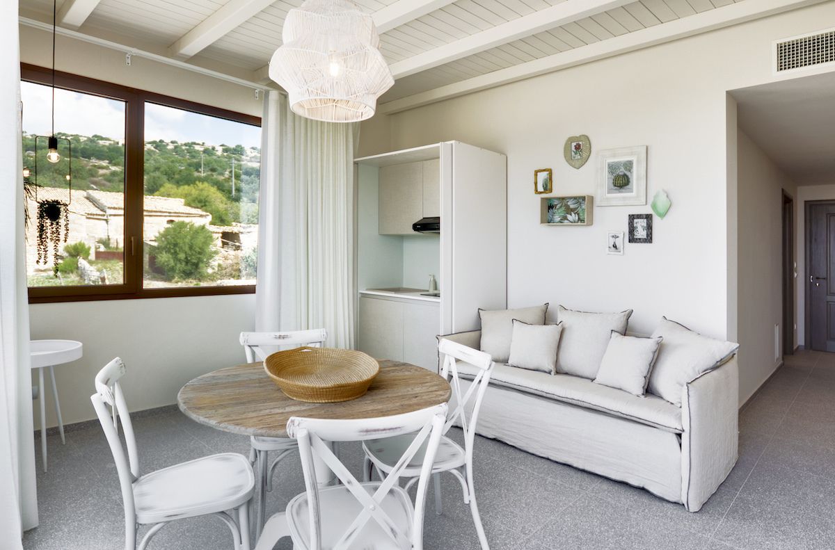 Una habitación moderna y luminosa con un sofá blanco, una mesa redonda de madera con sillas y un ventanal que ofrece vistas a zonas verdes y edificios.