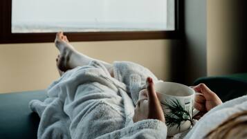 A person lying on a bed in a white bathrobe, holding a mug with a pine branch design.