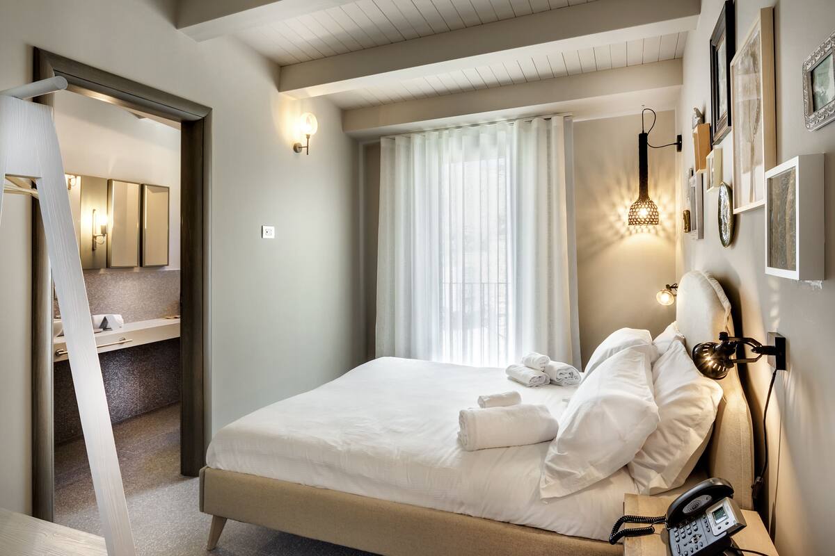 Habitación de hotel con una cama, un teléfono en la mesita de noche, una lámpara de pared y una ventana con cortinas.