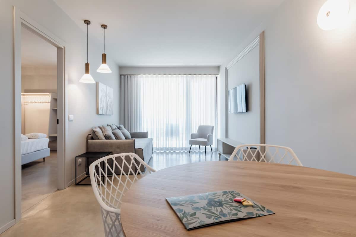 Una sala moderna con una mesa de comedor de madera, sillas blancas, un sofá, un televisor montado en la pared y un ventanal con cortinas translúcidas.