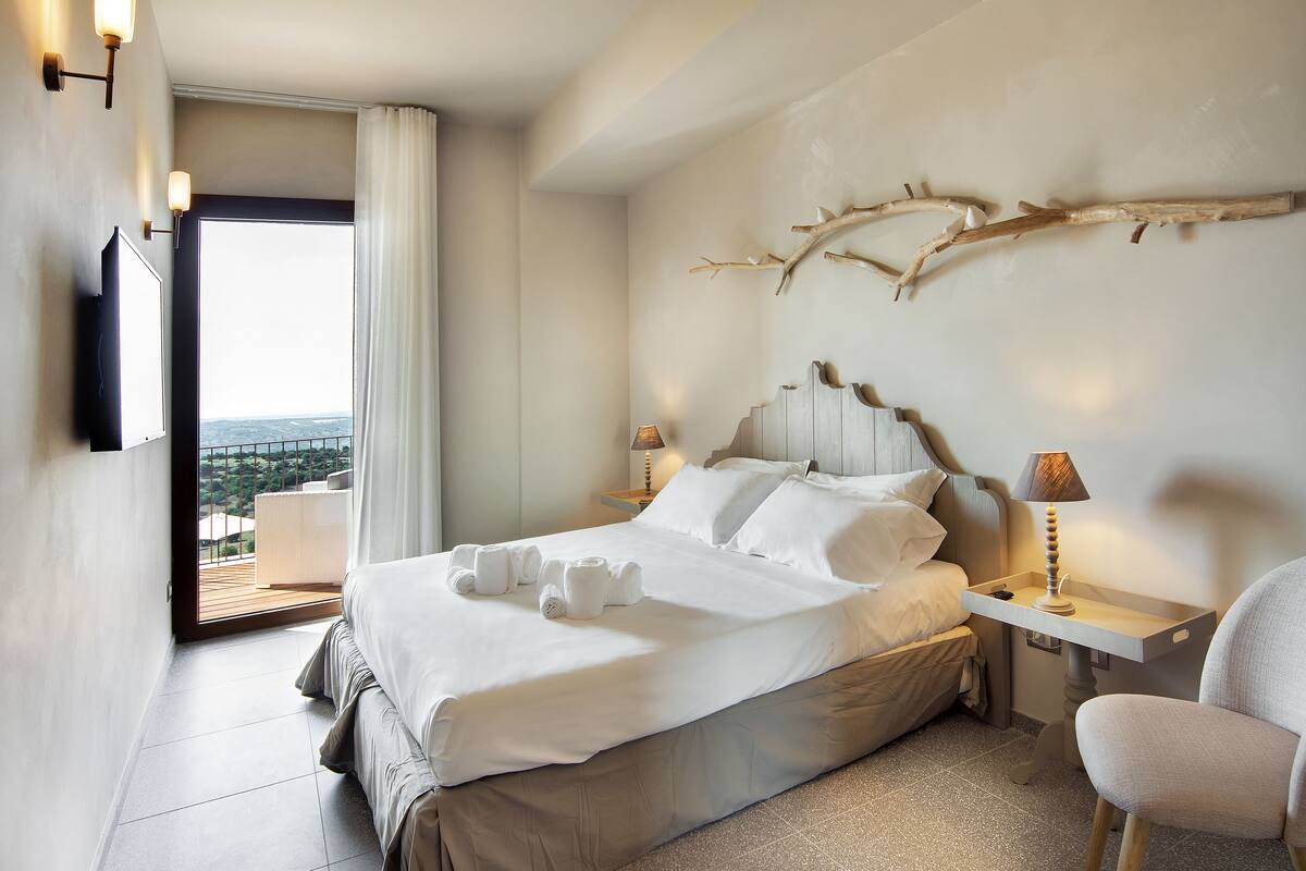 Habitación de hotel con una cama grande, una mesita de noche con lámpara, una silla y vistas al exterior.