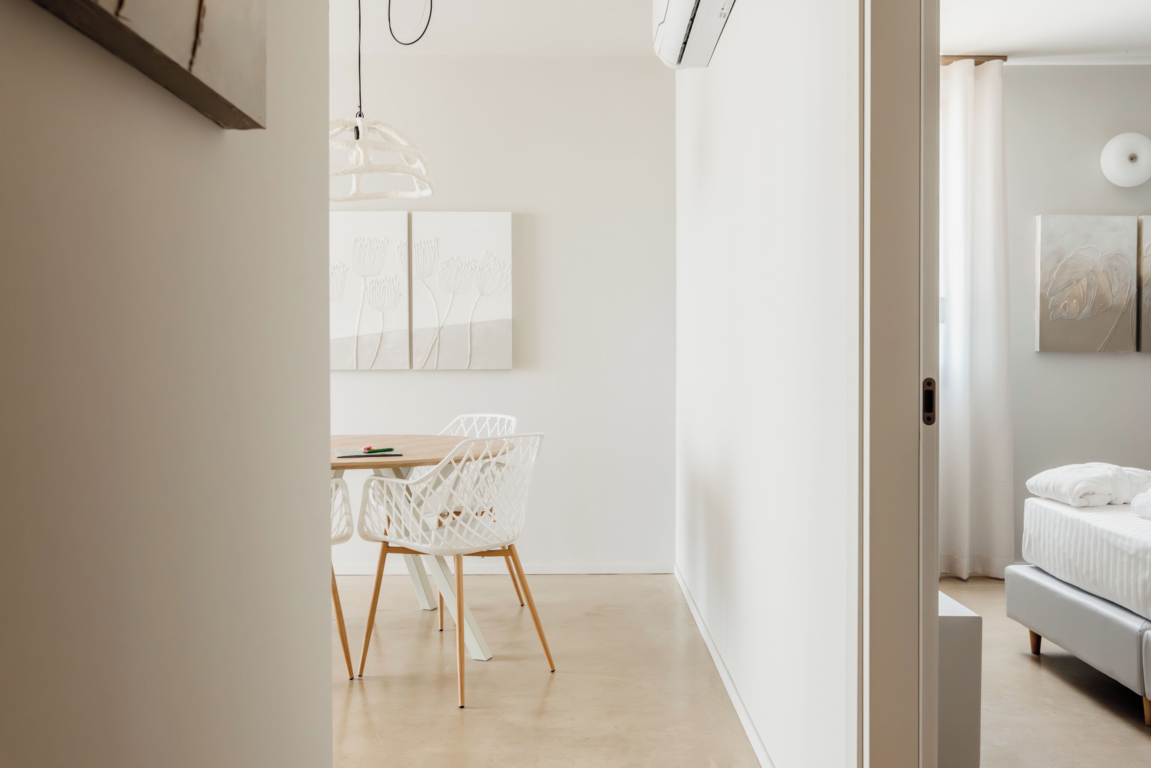 Een moderne, minimalistisch ingerichte kamer met een houten eettafel en witte stoelen, een hanglamp en een bed met witte beddengoed.