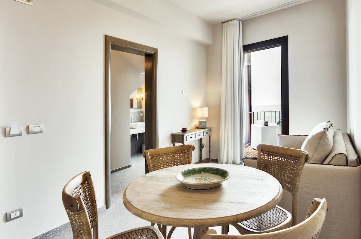 Une chambre d’hôtel avec une table à manger ronde, des chaises en osier, un canapé, une petite table d’appoint avec une lampe, et une vue sur l’extérieur par une fenêtre.