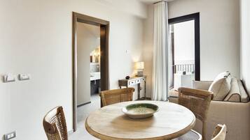 Une chambre d’hôtel avec une table à manger ronde, des chaises en osier, un canapé, une petite table d’appoint avec une lampe, et une vue sur l’extérieur par une fenêtre.