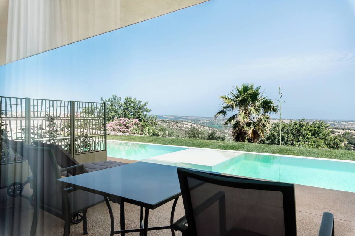 Un balcón con una mesa y sillas de vidrio, con vistas a una piscina y un paisaje que incluye colinas y una masa de agua.