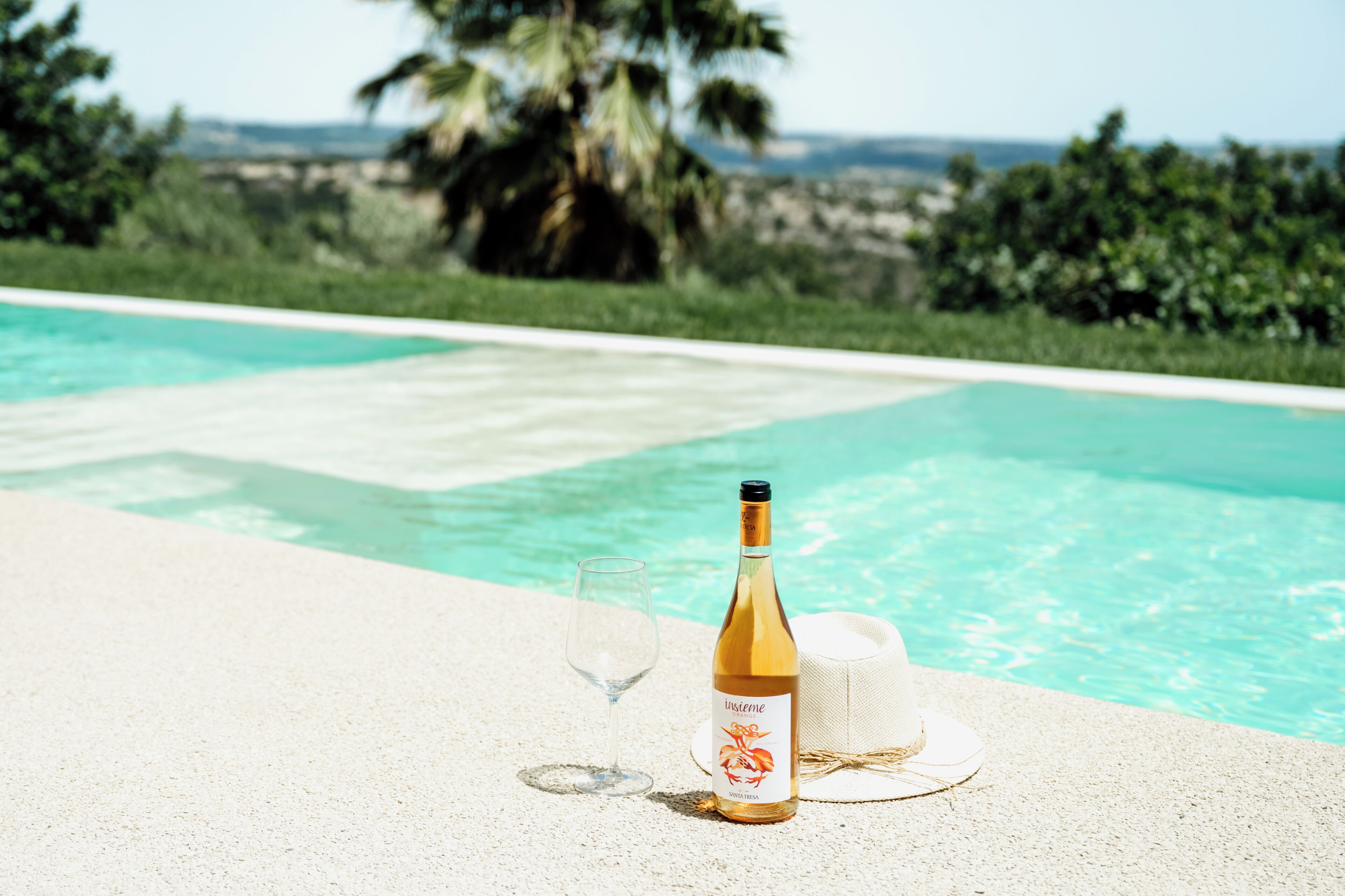 A bottle of wine and a glass on a poolside table with a hat, overlooking a pool with clear water and palm trees in the background.