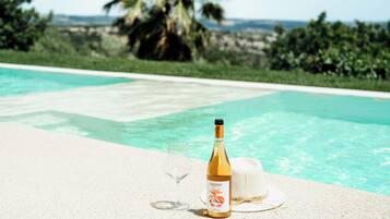 A bottle of wine and a glass on a poolside table with a hat, overlooking a pool with clear water and palm trees in the background.