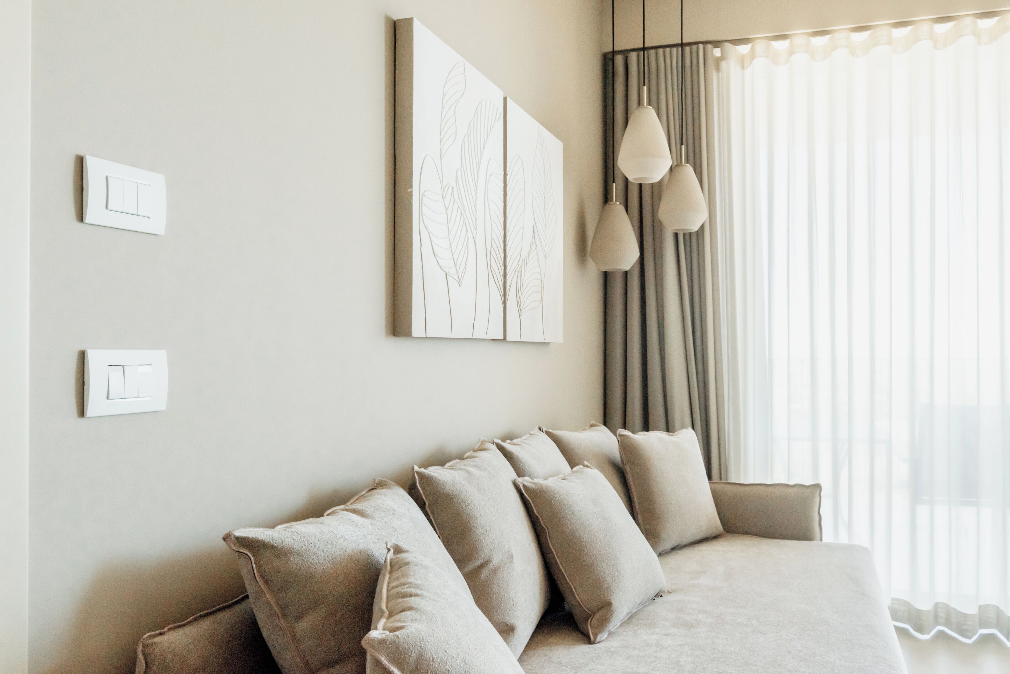 Quarto moderno com sofá bege, dois interruptores de luz na parede e uma janela grande com cortinas.