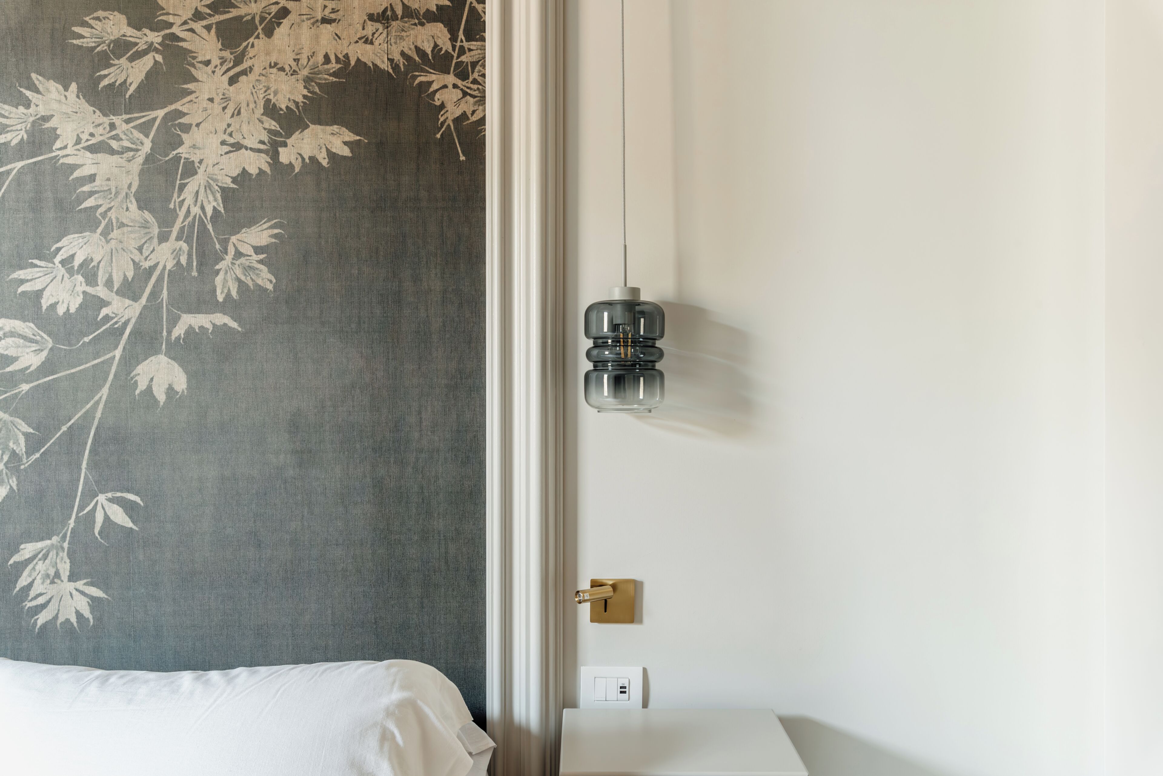 Une chambre d’hôtel comprenant un lit, une table de chevet, une lampe fixée au mur et un mur orné d’un motif de feuilles.