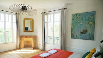 A bedroom with a bed, a fireplace, a large window, and a painting.