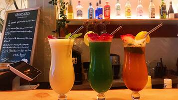 Trois cocktails colorés sont présentés sur le comptoir en bois d’un bar, avec une sélection de bouteilles de spiritueux en arrière-plan.